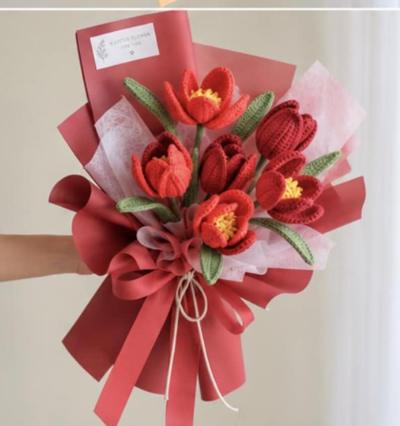 May include: A bouquet of crocheted red tulips with yellow centers and green leaves, wrapped in dark red and translucent white paper. A small white rectangular tag at the top features a floral illustration and the text A LITTLE FLOWER FOR YOU.