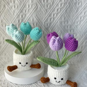 May include: Two crochet tulip arrangements in white pots with smiling faces and brown feet. One arrangement features blue-green tulips, the other purple. Green leaves and stems support the flowers, creating a whimsical, handmade aesthetic.