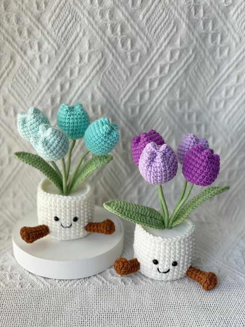 May include: Two crochet tulip arrangements in white pots with smiling faces and brown feet. One arrangement features blue-green tulips, the other purple. Green leaves and stems support the flowers, creating a whimsical, handmade aesthetic.