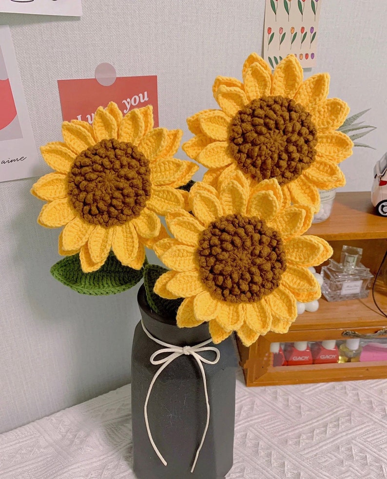 May include: Three crocheted sunflowers in a black vase with a white ribbon. The sunflowers are yellow with brown centers and green leaves.