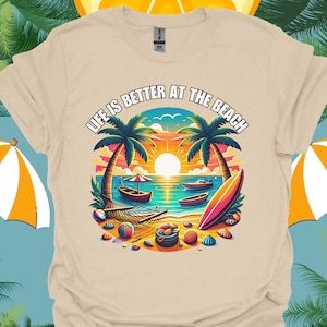 May include: A beige t-shirt with a colorful graphic design of a beach scene. The graphic includes palm trees, a hammock, boats, a surfboard, and the text "Life is better at the beach".