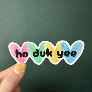 May include: A sticker with the words "ho duk yee" in black letters, each word on a heart-shaped design. The hearts are colored pink, green, yellow, and blue. The sticker is held against a dark green background.