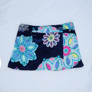 Puede incluir: Una falda azul marino oscuro con un vibrante estampado floral geométrico multicolor en tonos rosa, azul claro, amarillo y blanco. La falda presenta un diseño cruzado, una cinturilla con dos botones oscuros y una textura suave y aterciopelada.
