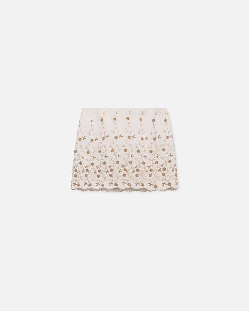 Puede incluir: Una falda color crema con intrincados bordados florales en un tono marr&oacute;n dorado. La falda presenta un dobladillo festoneado y un dise&ntilde;o delicado y femenino. El bordado crea un efecto texturizado, agregando inter&eacute;s visual a la prenda.