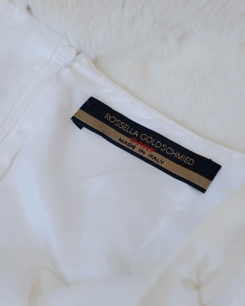 Puede incluir: Primer plano de una prenda blanca con cremallera y una etiqueta negra y dorada. La etiqueta dice "ROSSELLA GOLDSCHMIED" y "MADE IN ITALY". La prenda parece estar hecha de un material suave y sedoso.