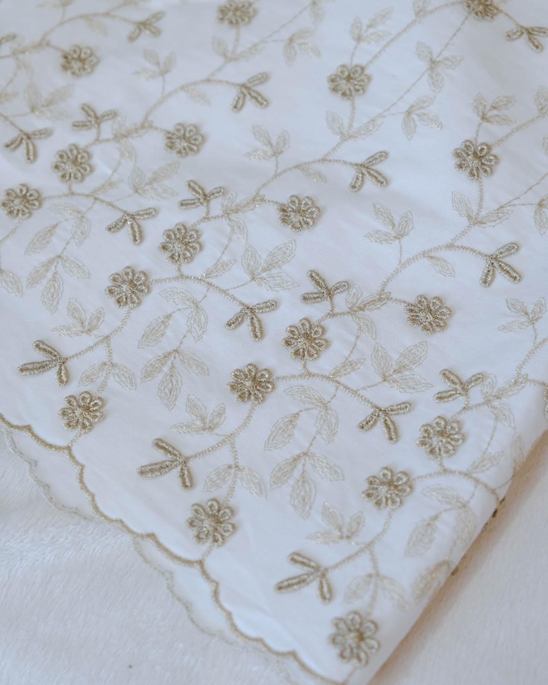 Puede incluir: Tela blanca con intrincados bordados dorados con motivos florales y de hojas. El dise&ntilde;o incluye delicadas flores y vides, creando un textil elegante. El borde de la tela tiene un ribete festoneado.