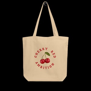 May include: A beige tote bag with a red circle design featuring two red cherries and the text "CHERRY RED AMBITION".