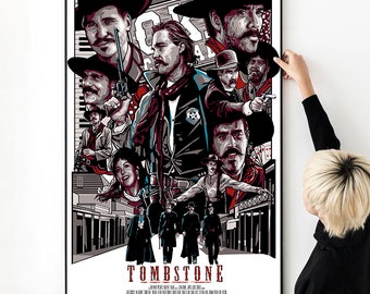 May include: A framed movie poster for the film "Tombstone" featuring a collage of characters in Western attire. The poster has a black border and is being held against a white wall. The poster's color palette includes red, black, white, and gray.