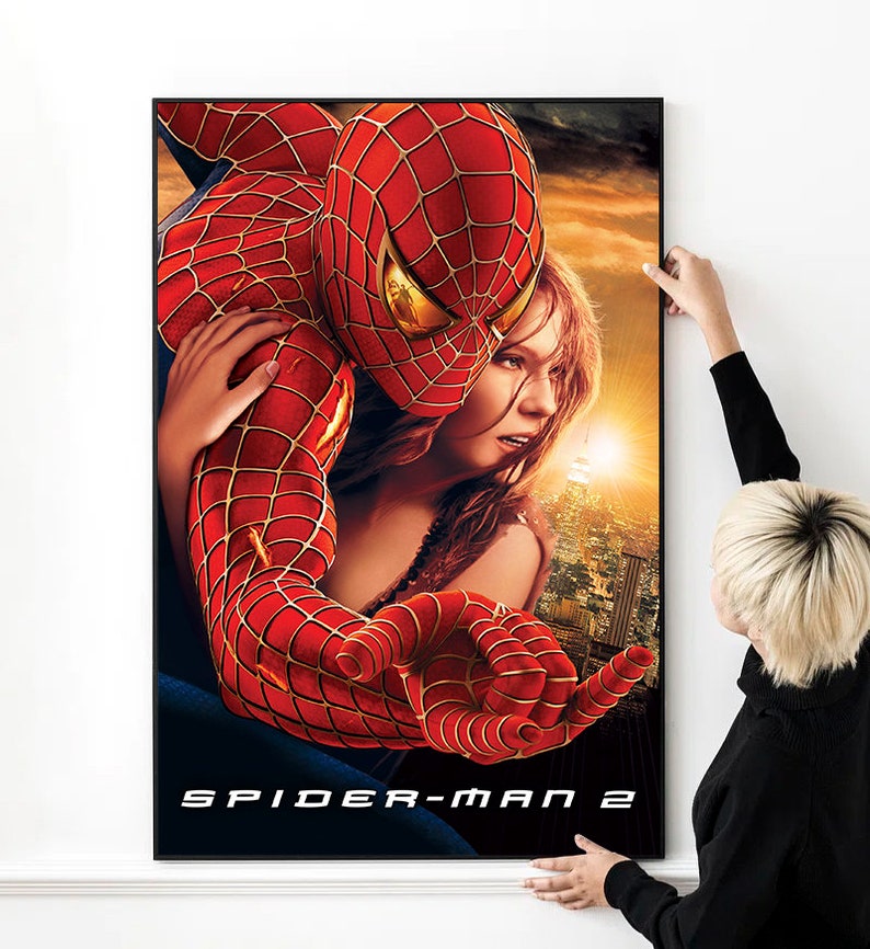 May include: A movie poster for Spider-Man 2 featuring Spider-Man in his red and blue suit holding a woman in a white tank top. The poster has a cityscape background with a sunset in the distance. The text "SPIDER-MAN 2" is at the bottom of the poster.