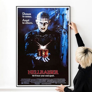 Puede incluir: Un póster de película de terror vintage para la película Hellraiser. El póster presenta un fondo azul oscuro con una figura amenazante que sostiene una caja de rompecabezas. El texto en el póster dice: "Demon to some. Angel to others. Hellraiser. He'll tear your soul apart."