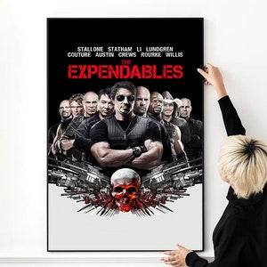 Op de afbeelding: Een ingelijste filmposter voor "The Expendables" met een zwarte achtergrond en een groep mannen. De titel is rood en een schedel- en wapenontwerp staat onderaan. Een persoon houdt de poster vast.