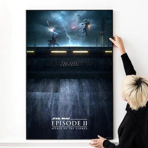 May include: A framed Star Wars Episode II: Attack of the Clones poster. The artwork depicts a battle scene with characters wielding lightsabers under a stormy sky. The text "THE SAGA CONTINUES" is above the title.