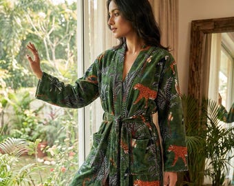 May include: A long, green robe with a jungle-themed pattern featuring orange leopards, trees, and birds. The robe has a tie waist, pockets, and a striped lining. The model is standing near a window.