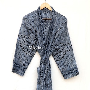 Può includere: Una lunga vestaglia fluida con un motivo paisley nei toni del blu e del nero. La vestaglia è annodata in vita e appesa a una gruccia di legno. La parola "Mishika" è stampata sulla vestaglia.
