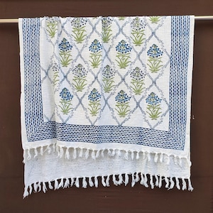 Puede incluir: Manta de algodón blanca con un estampado floral azul y ribete de flecos. El diseño presenta flores azules y hojas verdes dentro de un enrejado de diamantes, enmarcado por un borde azul. La manta está colgada de una barra de metal.