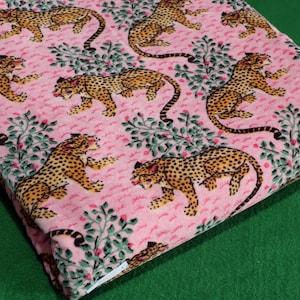 Può includere: Tessuto rosa con un motivo ripetuto di ghepardi e rami verdi con foglie. I ghepardi sono marroni con macchie nere. Il tessuto è piegato, rivelando il motivo su una superficie verde.