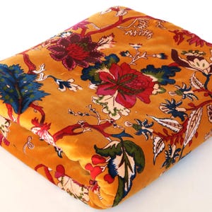 Puede incluir: Tela doblada de color mostaza con un estampado floral. El diseño presenta grandes flores rojas, azules y verdes con detalles en blanco y rojo. La tela está doblada cuidadosamente, mostrando los intrincados detalles del estampado.