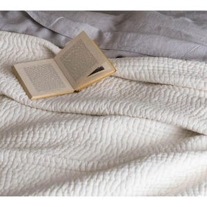 Puede incluir: Un edredón acolchado de color crema con un patrón texturizado. Un libro antiguo con páginas amarillentas está abierto encima del edredón. Una sábana gris es visible en el fondo, creando una paleta de colores neutros.