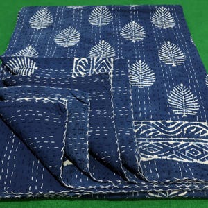 Op de afbeelding: Een gevouwen indigo blauwe kantha quilt met witte bladeren patronen en decoratieve rand. De quilt is met de hand gestikt met witte draad, een traditioneel textielontwerp. De achtergrond is effen groen.