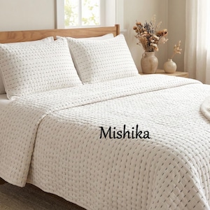 Op de afbeelding: Een wit sprei en twee bijpassende kussenslopen met een gestikt patroon worden getoond op een houten bedframe. Het woord "Mishika" staat op het sprei gedrukt. Een beige deken is over het bed gedrapeerd.