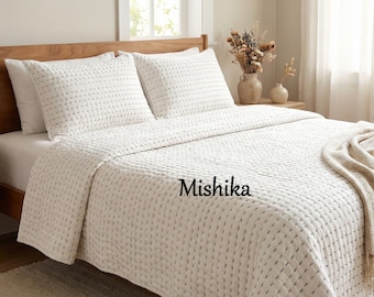 May include: A white bedspread and two matching pillowcases with a stitched pattern are displayed on a wooden bed frame. The word "Mishika" is printed on the bedspread. A beige throw blanket is draped over the bed.