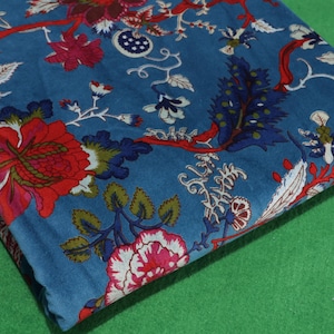 Puede incluir: Tela azul doblada con un estampado floral. El diseño presenta grandes flores rojas y rosas, hojas verdes y detalles blancos sobre un fondo azul. Ramas y tallos rojos conectan los elementos florales.