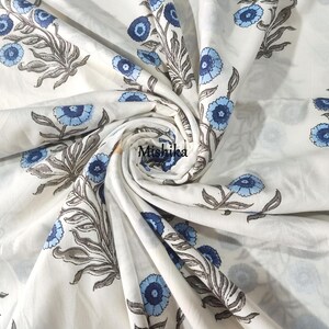 Puede incluir: Tela blanca con un estampado floral en tonos azules y grises. El diseño presenta flores y hojas estilizadas, creando una estética delicada. La tela está doblada, mostrando la textura y la caída del material. La palabra "Mishika" es visible.