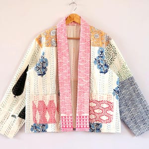 Op de afbeelding: Een jas met lange mouwen en een patchworkontwerp met diverse bloem- en geometrische patronen in tinten roze, blauw, geel en zwart op een crèmekleurige achtergrond. Het kledingstuk heeft een brede kraag, twee voorzakken en zichtbare stiksels.