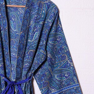 Op de afbeelding: Een kamerjas met lange mouwen voorzien van een levendig blauw paisley-patroon met ingewikkelde krullen in tinten blauwgroen, marineblauw en goud. Het kledingstuk heeft een bijpassende stoffen ceintuur in de taille en een brede rand aan de mouwboorden.