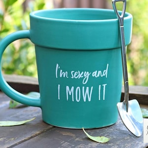 Peut inclure: Mug en céramique turquoise avec le texte "I'm sexy and I MOW IT" en écriture blanche. Une petite pelle argentée est posée contre la tasse. La tasse est sur une surface en bois.