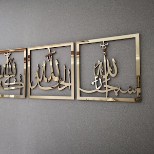 May include: Three gold-colored framed wall art pieces with Arabic calligraphy. Each piece features a different script within a square frame. The art is mounted on a gray textured wall, creating a contrast between the shiny gold and the matte background.