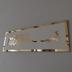 Op de afbeelding: Een gouden metalen wandkunstwerk met Arabische kalligrafie in een wit frame. De kalligrafie luidt "Bismillah Al-Rahman Al-Rahim", wat vertaald wordt als "In de naam van Allah, de Meest Barmhartige, de Meest Genadevolle."