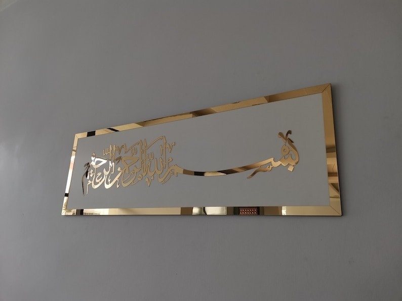 Op de afbeelding: Een gouden metalen wandkunstwerk met Arabische kalligrafie in een wit frame. De kalligrafie luidt "Bismillah Al-Rahman Al-Rahim", wat vertaald wordt als "In de naam van Allah, de Meest Barmhartige, de Meest Genadevolle."