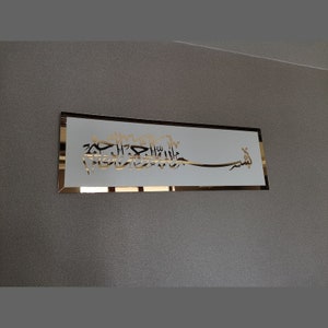 Op de afbeelding: Een rechthoekige witte plaquette met een gouden rand en gouden Arabische kalligrafie. De kalligrafie luidt "Bismillah Al-Rahman Al-Rahim", wat vertaald wordt als "In de naam van Allah, de Meest Barmhartige, de Meest Genadevolle."