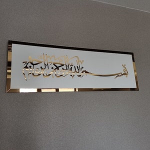 Op de afbeelding: Een gouden metalen wanddecoratie met Arabische kalligrafie in een wit frame. De kalligrafie luidt "Bismillah Al-Rahman Al-Rahim".