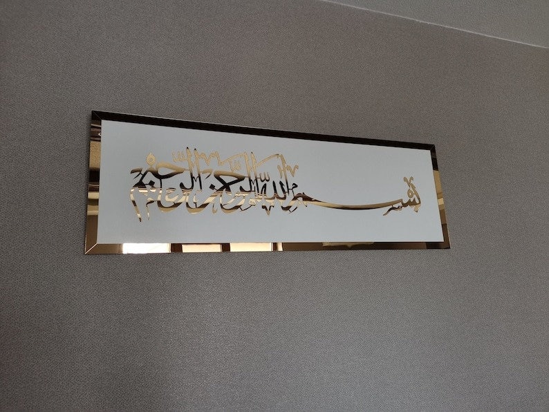 Op de afbeelding: Een gouden metalen wanddecoratie met Arabische kalligrafie in een wit frame. De kalligrafie luidt "Bismillah Al-Rahman Al-Rahim".