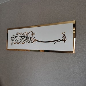 Op de afbeelding: Een gouden ingelijst wandkunstwerk met een witte achtergrond met Arabische kalligrafie in zwart. De kalligrafie luidt "Bismillah Al Rahman Al Rahim", wat vertaald wordt als "In de naam van Allah, de Meest Barmhartige, de Meest Genadevolle."