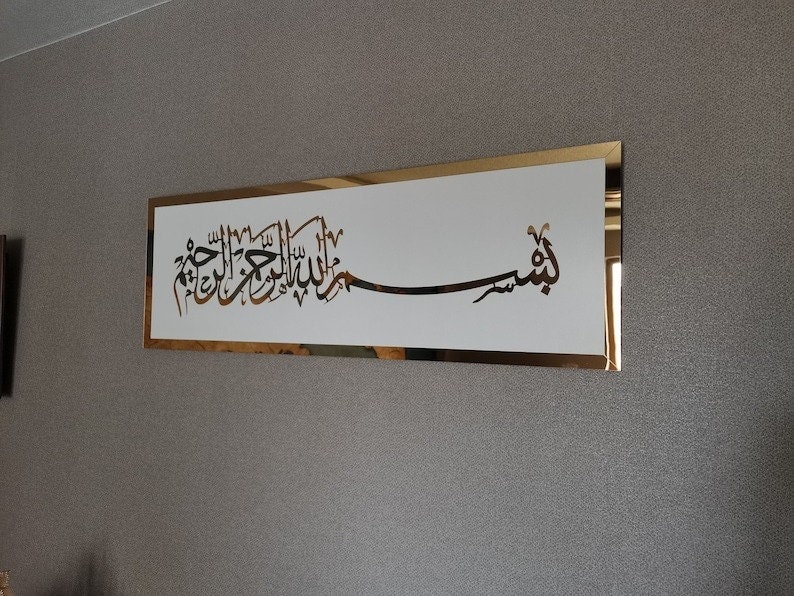 Op de afbeelding: Een gouden ingelijst wandkunstwerk met een witte achtergrond met Arabische kalligrafie in zwart. De kalligrafie luidt "Bismillah Al Rahman Al Rahim", wat vertaald wordt als "In de naam van Allah, de Meest Barmhartige, de Meest Genadevolle."