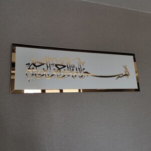 Op de afbeelding: Een gouden metalen plaquette met een witte achtergrond en Arabische kalligrafie in een decoratief lettertype. De tekst luidt "Bismillah Al-Rahman Al-Rahim".