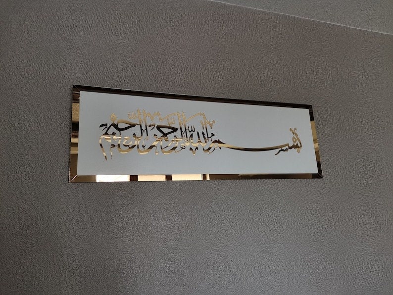 Op de afbeelding: Een gouden metalen plaquette met een witte achtergrond en Arabische kalligrafie in een decoratief lettertype. De tekst luidt "Bismillah Al-Rahman Al-Rahim".