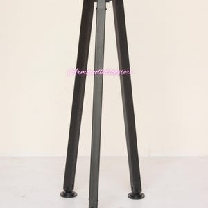 May include: A black metal tripod stand with three legs. The stand has a round top and is designed to hold an object. The legs are angled and have a decorative design at the base.