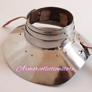Könnte beinhalten: Ein silberner Nackenschutz aus Metall mit einem Lederriemen. Der Nackenschutz hat eine abgerundete, ausgestellte Form und ist mit Nieten verziert. Der Lederriemen ist mit einer Schnalle am Nackenschutz befestigt.