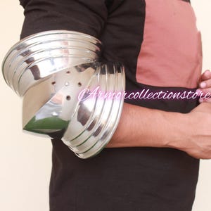 Puede incluir: Una protección de codo de metal plateado con un diseño en capas. La protección se lleva en el brazo izquierdo de una persona.