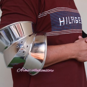 Puede incluir: Una protección de codo de metal plateado con un diseño redondeado y acampanado. La protección se lleva en el brazo izquierdo de una persona que lleva una camisa a rayas rojas y azules.