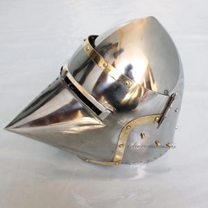 May include: A silver helmet with a pointed visor and brass accents. The helmet has a hinged visor and a brass strap on the side.
