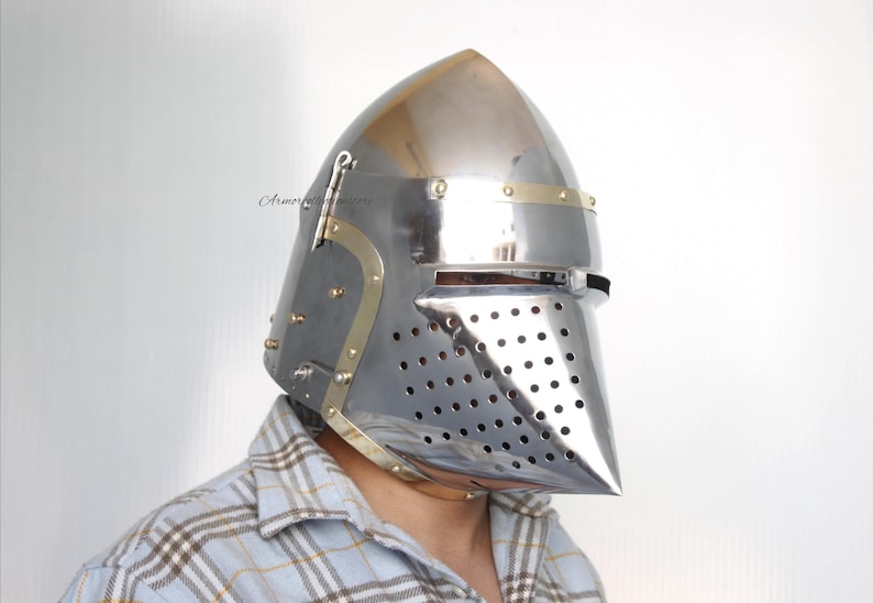 May include: A silver helmet with a pointed top and a face mask with a mesh design. The helmet has a brass trim around the edges and a brass band across the top.