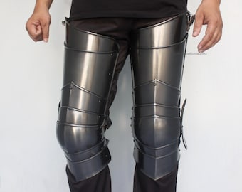 Puede incluir: Un par de armaduras de piernas de metal gris oscuro, con placas articuladas para mayor flexibilidad y protección. La armadura está diseñada para cubrir los muslos y las espinillas, con correas y hebillas para una sujeción segura. El metal tiene una superficie pulida y reflectante.