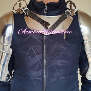 Könnte beinhalten: Ein Paar silberplattierte Schulterpanzer aus Stahl mit einem Lederriemen und einer Schnalle. Die Rüstung wird über einer schwarzen Tarnjacke getragen.