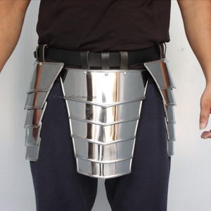 Puede incluir: Una pieza de armadura de cadera de metal plateado con placas superpuestas, usada sobre pantalones azul oscuro y un cinturón negro. La armadura tiene una superficie reflectante y está diseñada para proteger la sección media. La imagen muestra la armadura de la cintura hacia abajo.