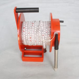May include: An orange and black measuring tape reel with a white tape marked in meters. The reel has a black handle and a metal plumb bob. The tape is rolled up inside the reel.